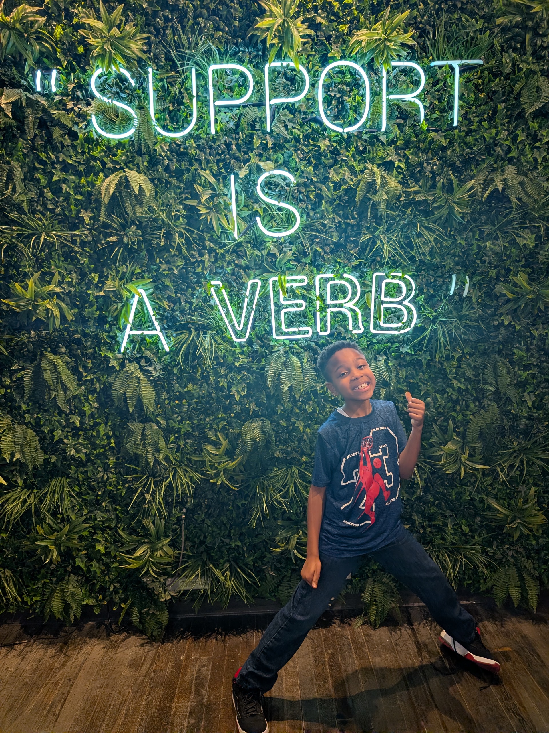 Shawntà's son Amir in front of a neon sign that reads Support is a verb